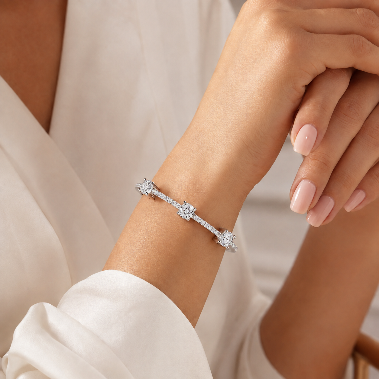 A close-up of a delicate silver-toned bracelet with three star-shaped diamond clusters, worn on a woman's wrist. The person is wearing a soft, off-white blouse, and her manicured hands are resting above her arm.