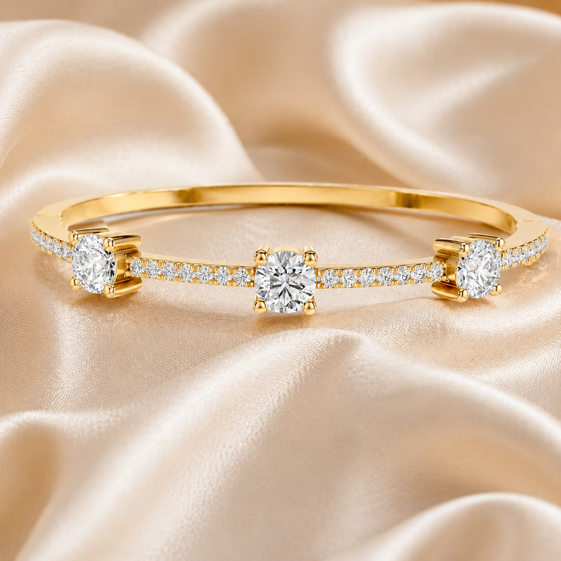 A yellow gold bangle bracelet with three round-cut center stones and pavé-set accent stones, displayed on draped champagne satin fabric.
