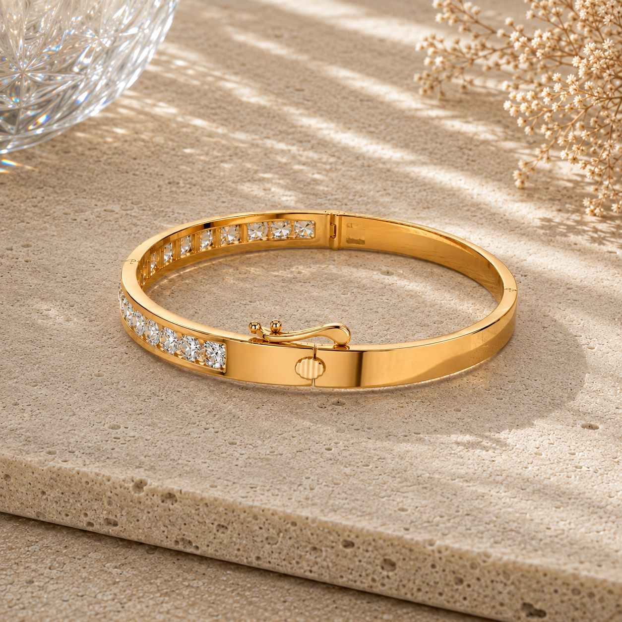 A polished yellow gold bangle bracelet with rows of small clear stones on the front, displayed on a textured beige stone surface with soft shadows and decorative dried flowers in the background.