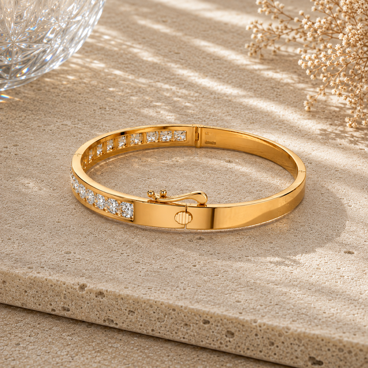 A polished yellow gold bangle bracelet with rows of small clear stones on the front, displayed on a textured beige stone surface with soft shadows and decorative dried flowers in the background.