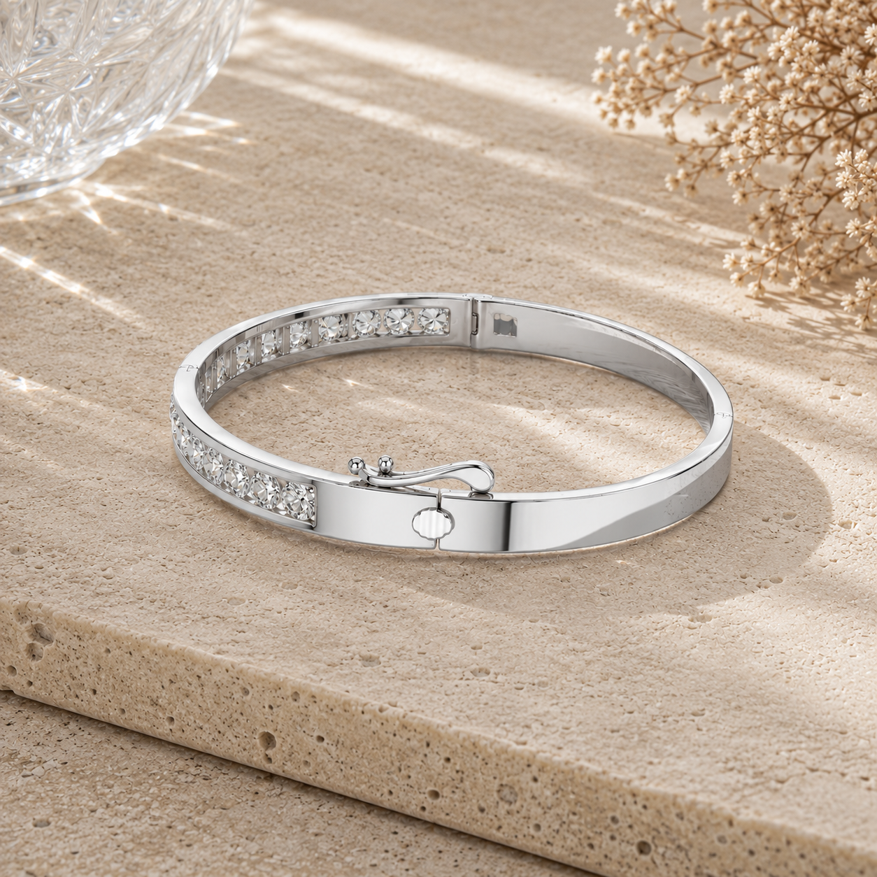A polished silver bangle bracelet with a row of clear stones on one side and a small central charm, displayed on a textured beige stone surface with dried flowers in the background.