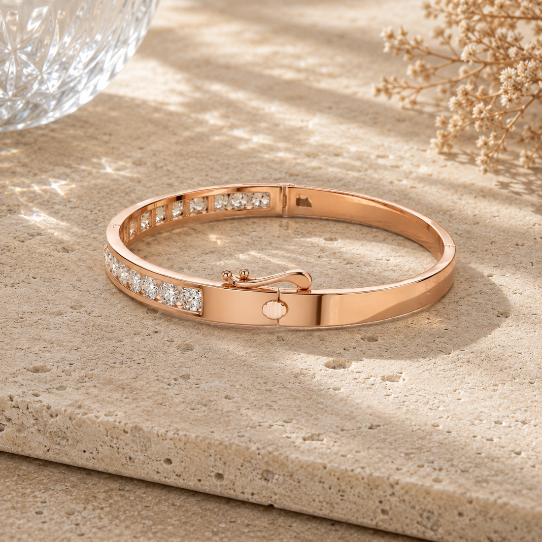 A rose gold bangle bracelet with a row of clear pavé-set stones on one side and a small round stone on the clasp, displayed on a textured beige stone surface with soft shadows.