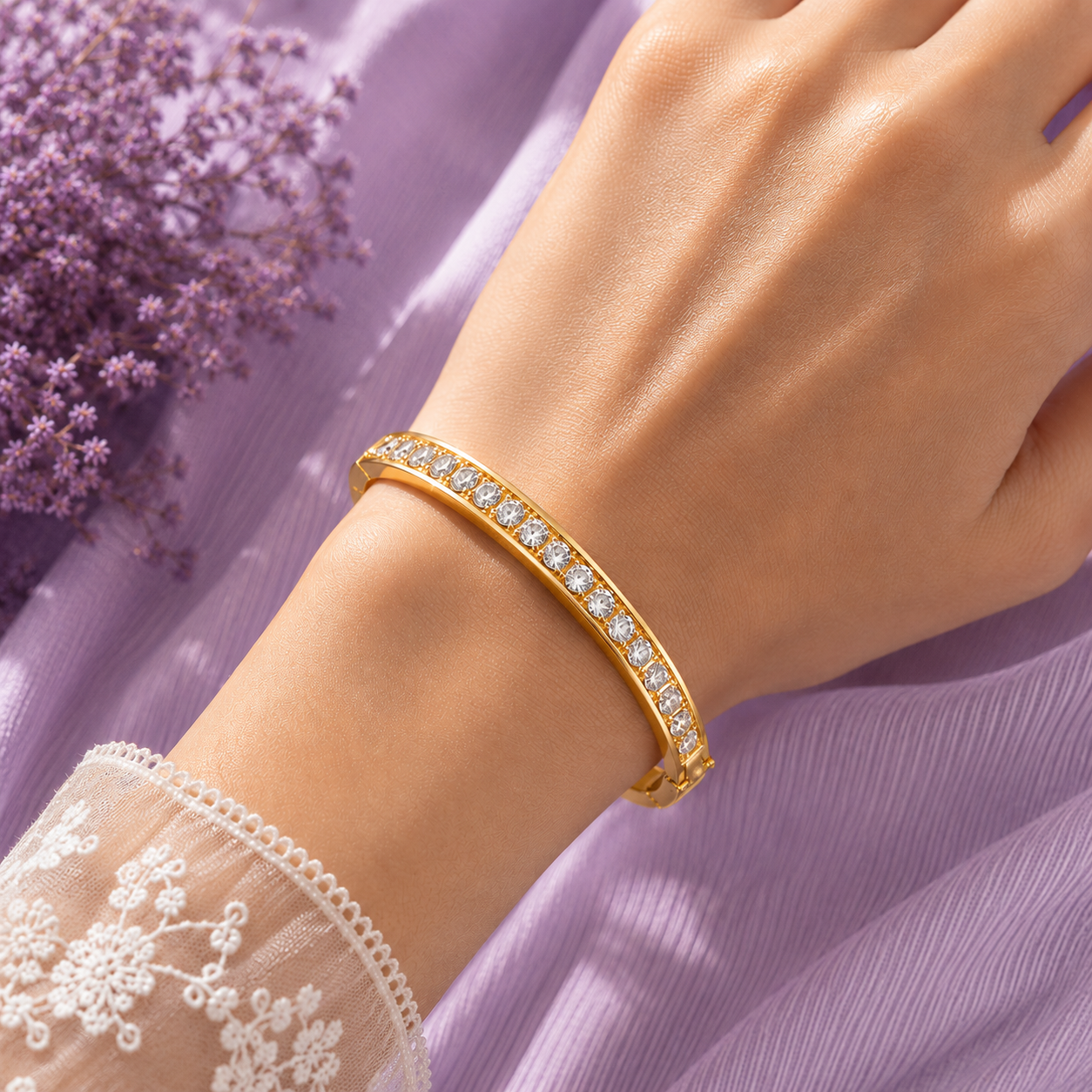 A gold bangle bracelet with a row of clear pavé-set stones, shown worn on a person's wrist against a soft lavender fabric background with purple flowers.
