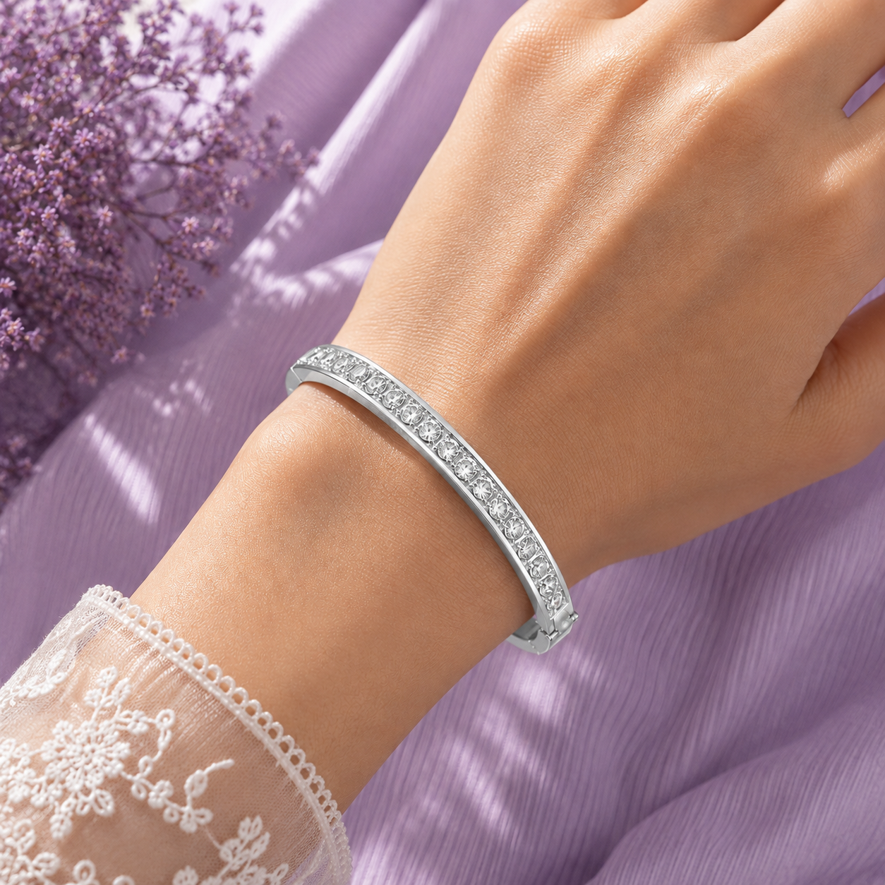 A close-up of a woman's wrist wearing a slim silver bangle bracelet with a row of clear stones, set against a soft lavender fabric background with a lace sleeve visible at the lower left.