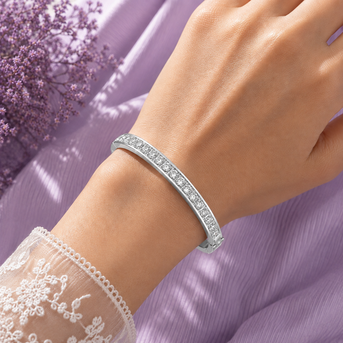 A close-up of a woman's wrist wearing a slim silver bangle bracelet with a row of clear stones, set against a soft lavender fabric background with a lace sleeve visible at the lower left.