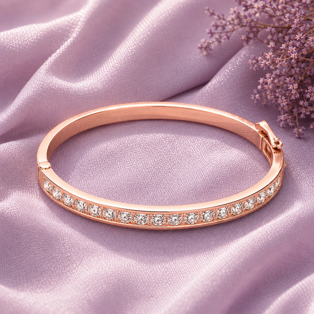 A rose gold bangle bracelet with a row of clear pavé stones, displayed on draped light purple satin fabric with small purple flowers in the corner.