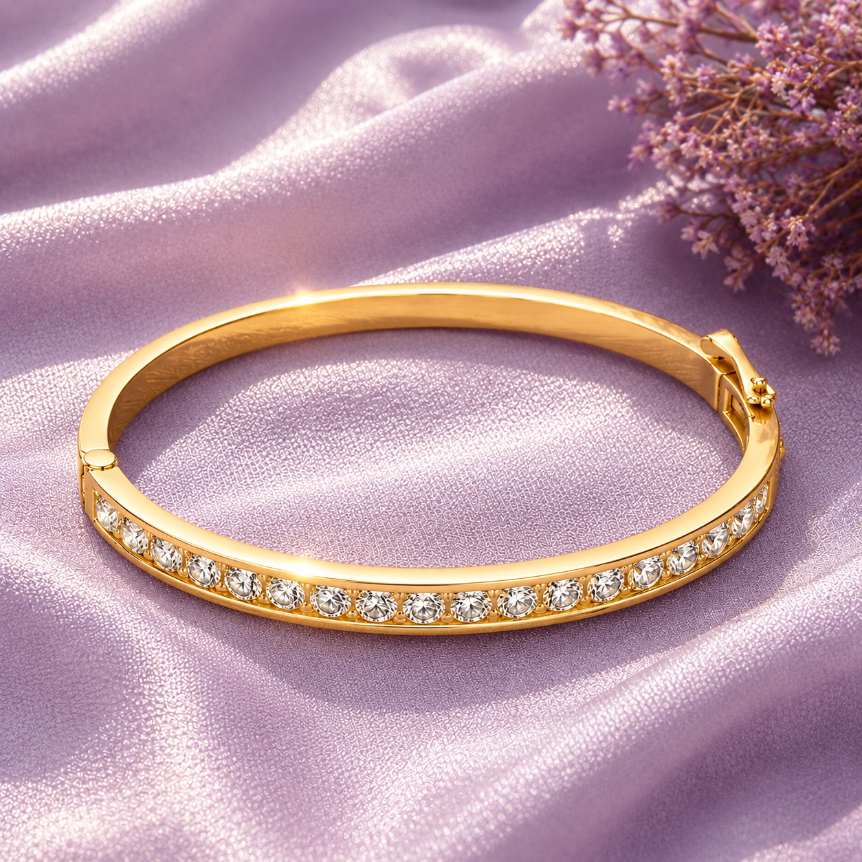 A gold bangle bracelet with a row of clear stones along the front, displayed on draped lavender satin fabric with small dried flowers in the corner.