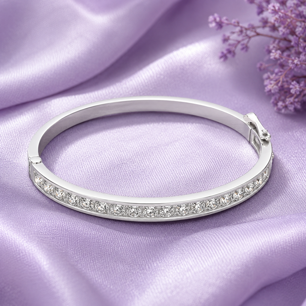 A polished silver bangle bracelet with a row of clear stones along the front, displayed on draped lavender satin fabric with small purple flowers in the corner.