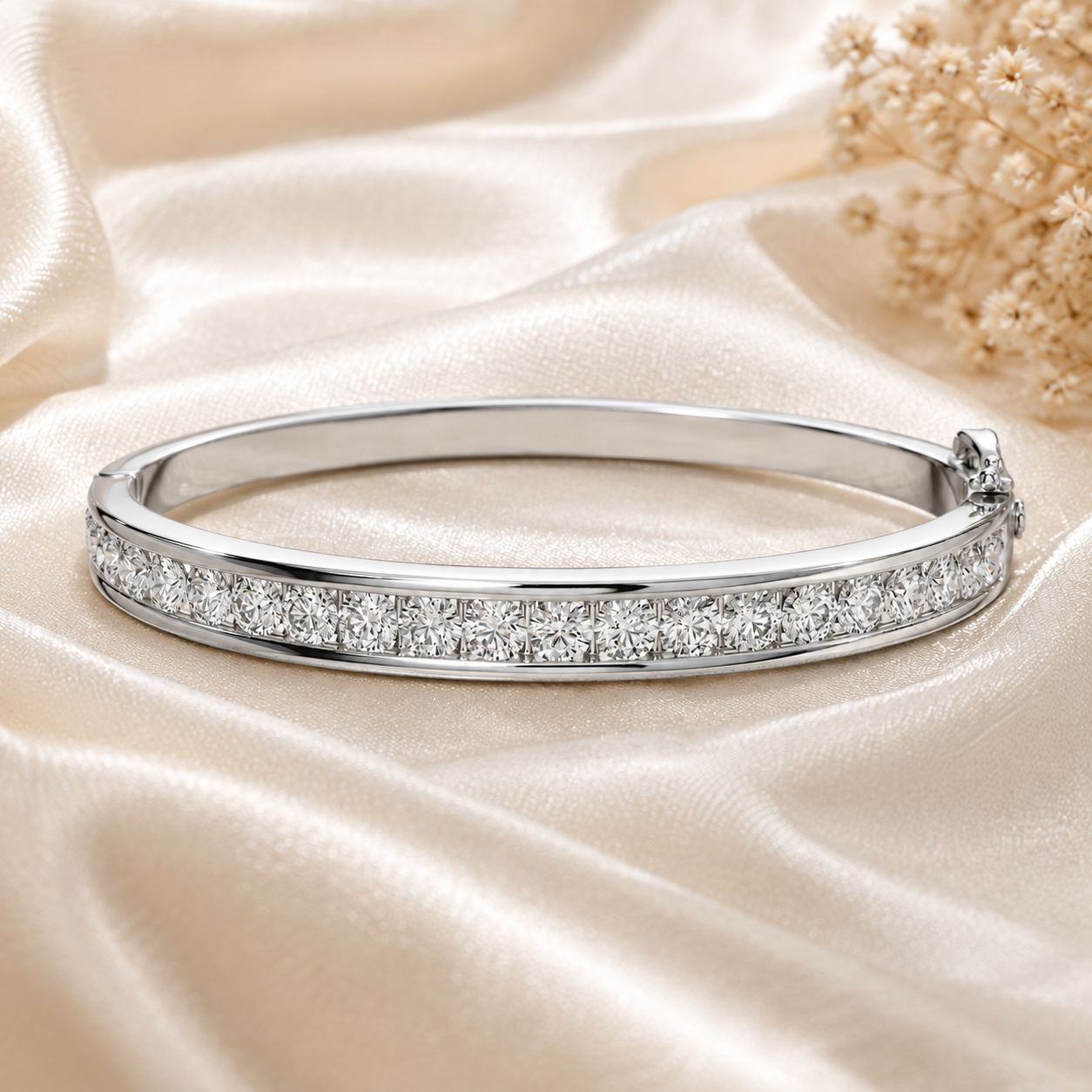 A polished silver-toned bangle bracelet paved with clear stones, displayed on draped champagne satin fabric with dried floral decor in the background.