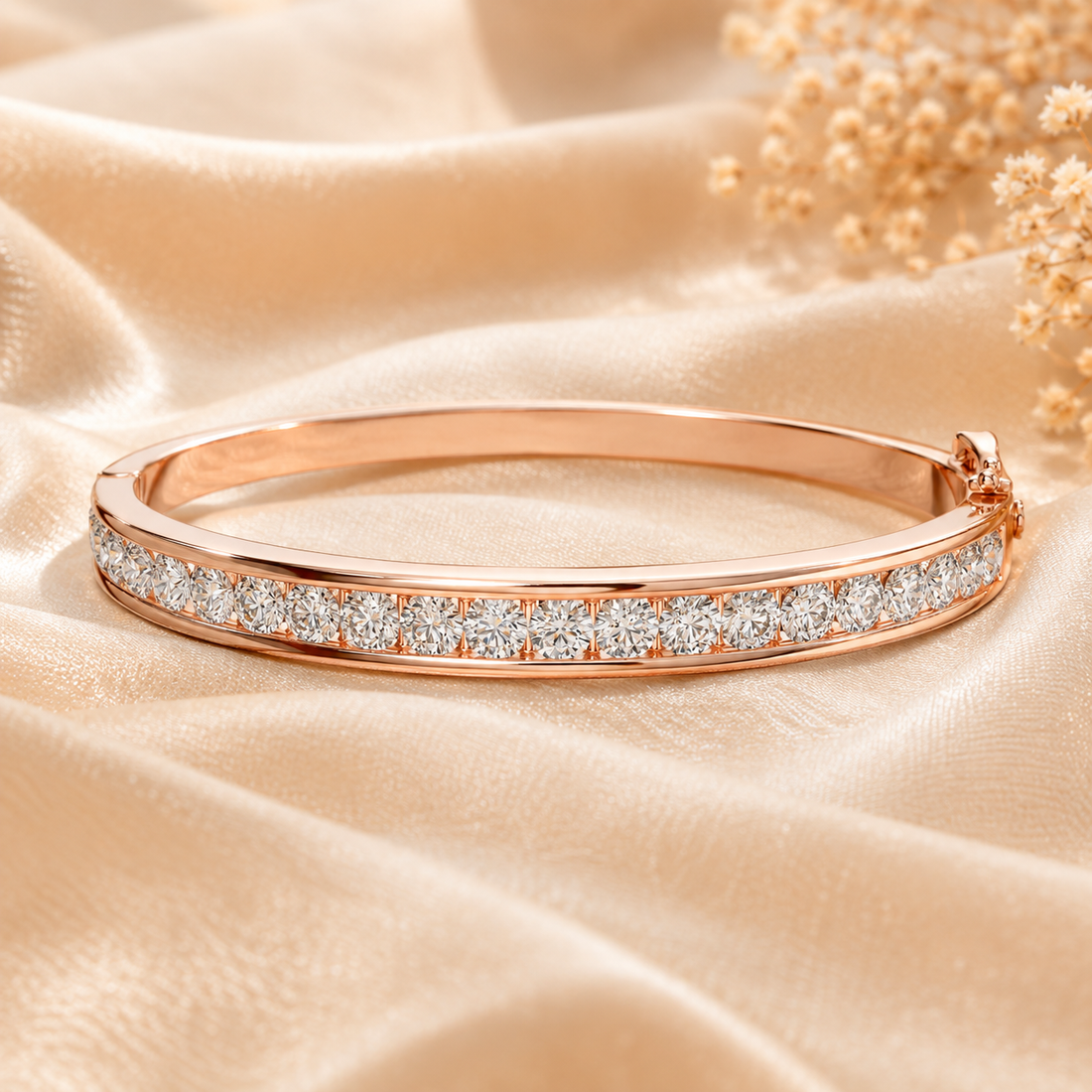A rose gold bangle bracelet with a channel-set row of clear stones, displayed on draped champagne-colored satin fabric with dried floral accents.
