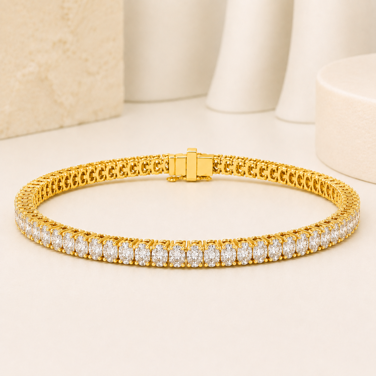 A gold tennis bracelet with a row of clear stones on the front, displayed on a light beige surface with soft, neutral background props.