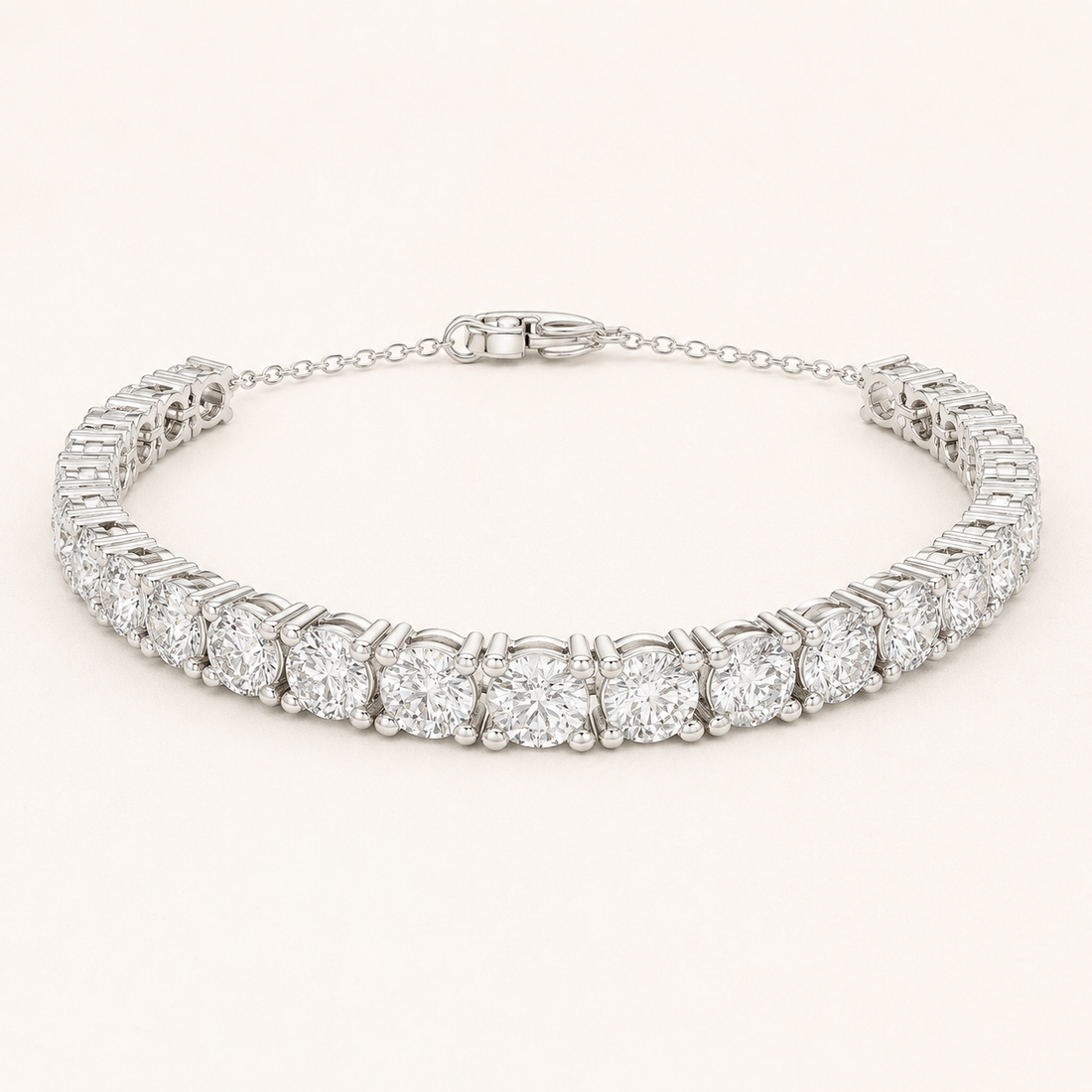 A diamond tennis bracelet in a silver-toned setting, featuring round-cut diamonds in a linked design with a delicate chain extender and clasp, shown on a light cream background.