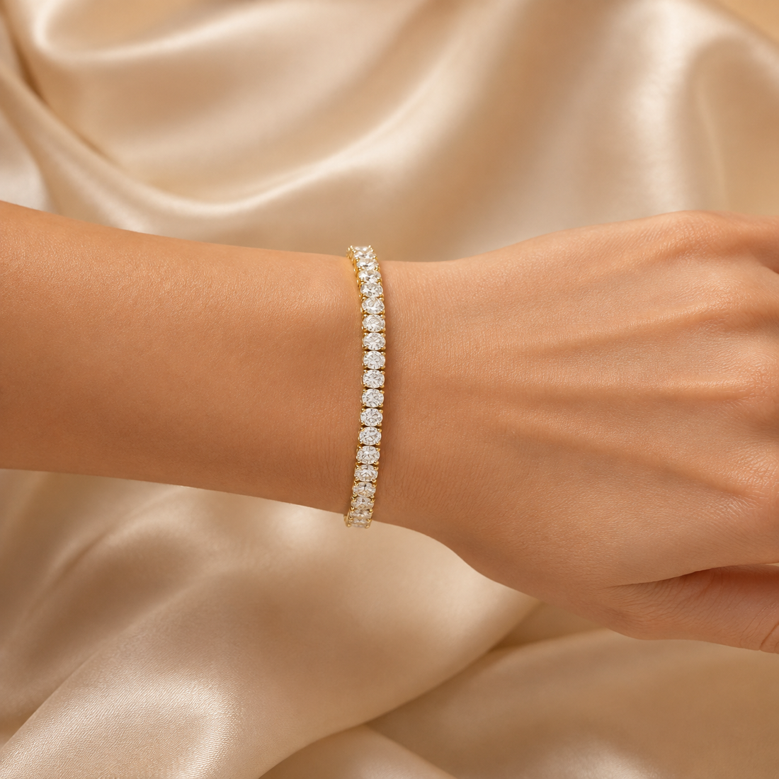 A close-up of a delicate tennis bracelet with round-cut clear stones set in yellow gold, worn on a person's wrist against a draped champagne satin fabric background.