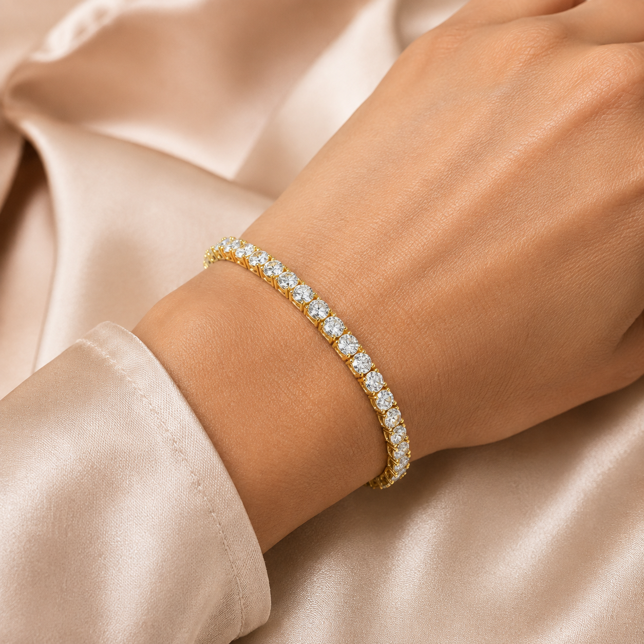 A gold tennis bracelet with round-cut clear stones worn on a woman's wrist, set against a soft champagne satin fabric background.