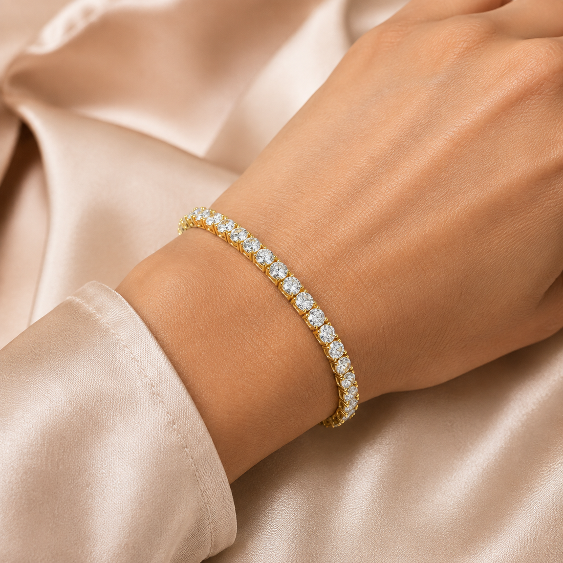 A gold tennis bracelet with round-cut clear stones worn on a woman's wrist, set against a soft champagne satin fabric background.