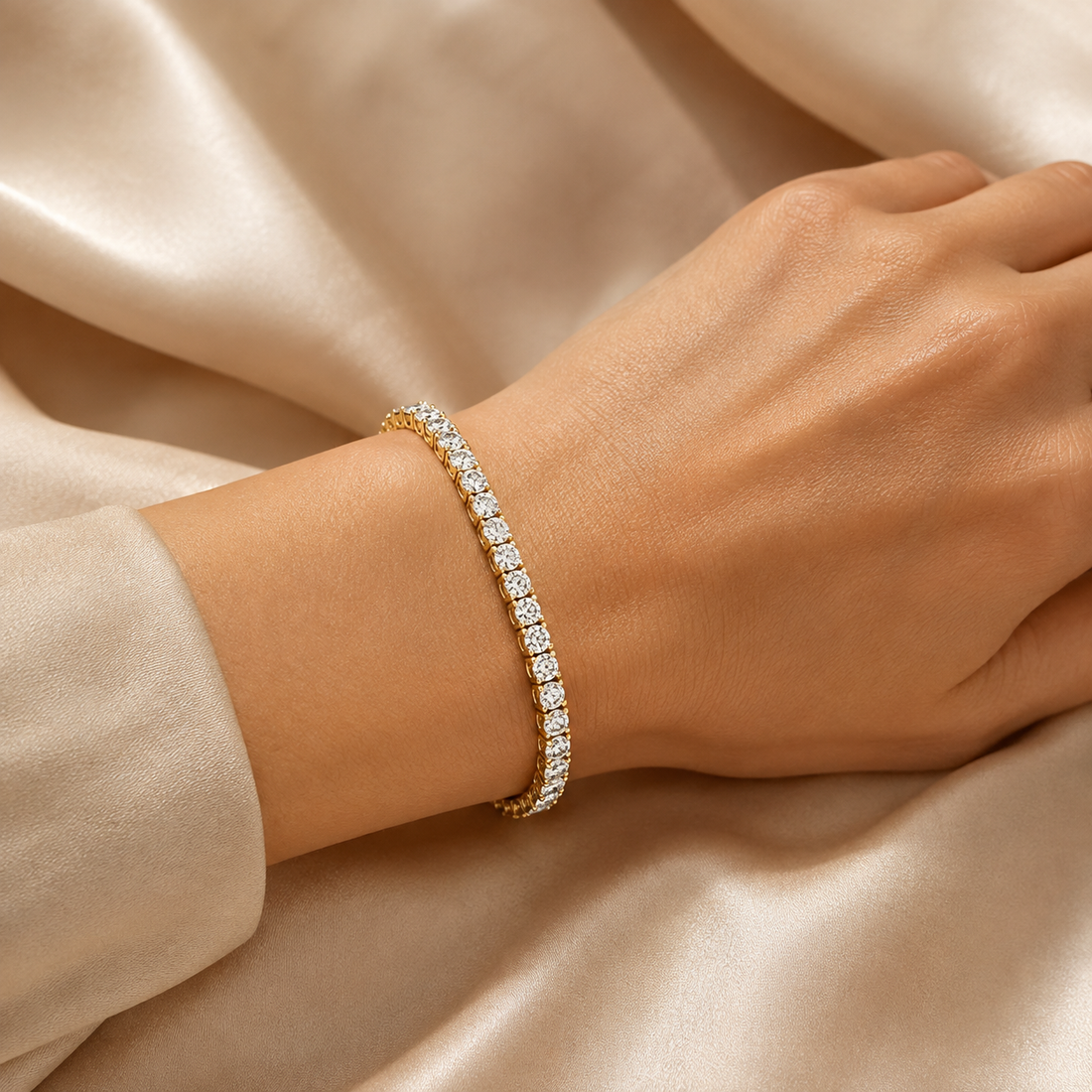 A close-up of a gold tennis bracelet with clear stones worn on a woman's wrist, set against a background of draped champagne-colored satin fabric.