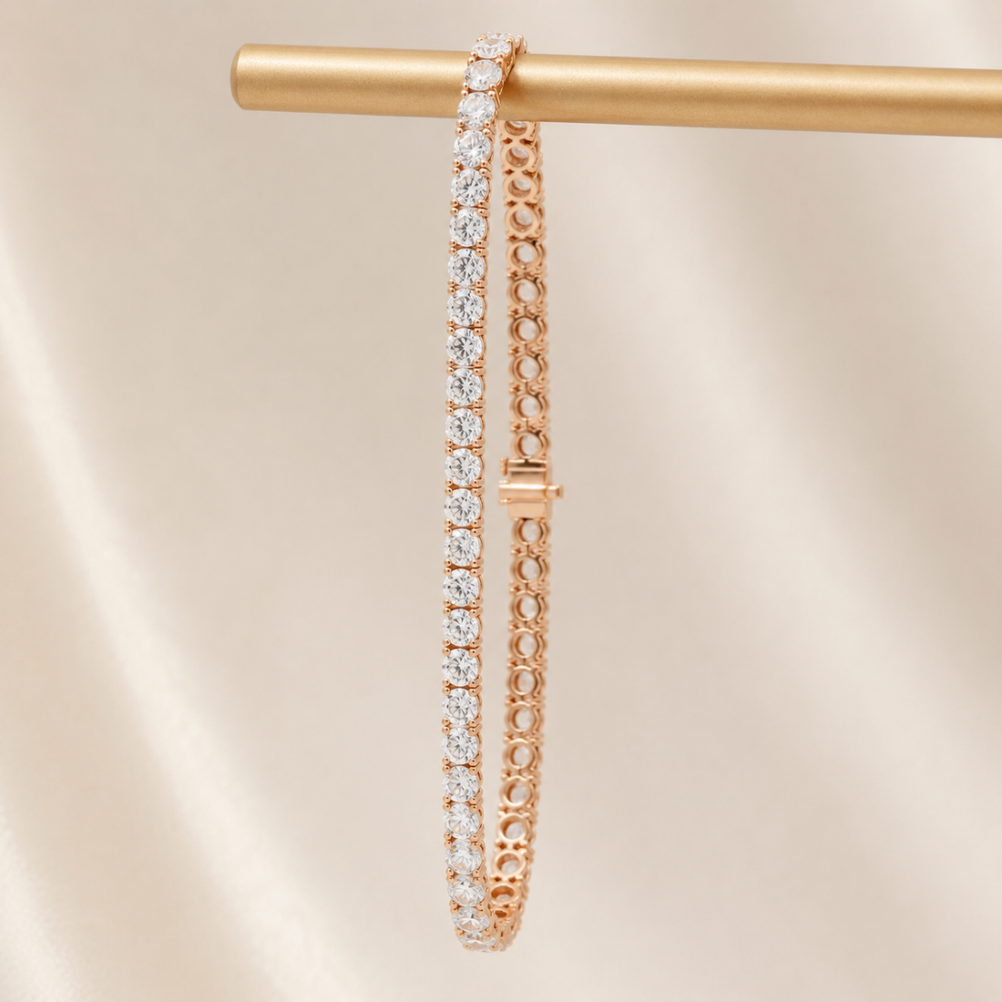 A rose gold tennis bracelet with sparkling clear stones, hanging from a gold display rod against a soft beige satin background.
