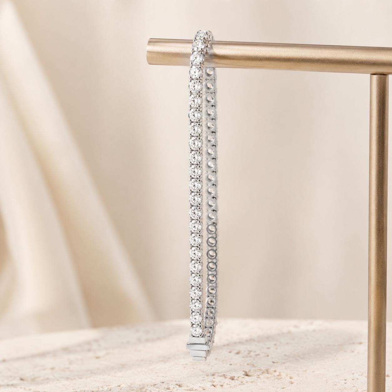 A silver tennis bracelet with clear round-cut stones, hanging from a gold display stand against a soft beige fabric background.