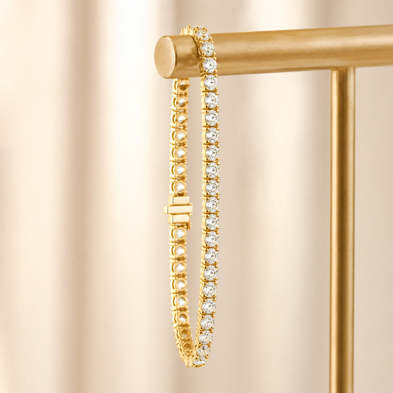 A gold tennis bracelet with clear round-cut stones, hanging from a gold display stand against a soft beige background.