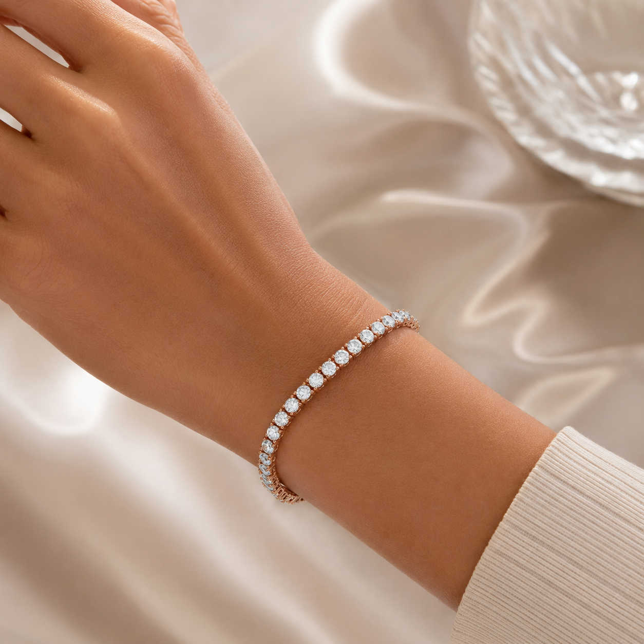 A close-up of a person's wrist wearing a delicate rose gold tennis bracelet with sparkling clear gemstones, set against a soft beige satin fabric background.