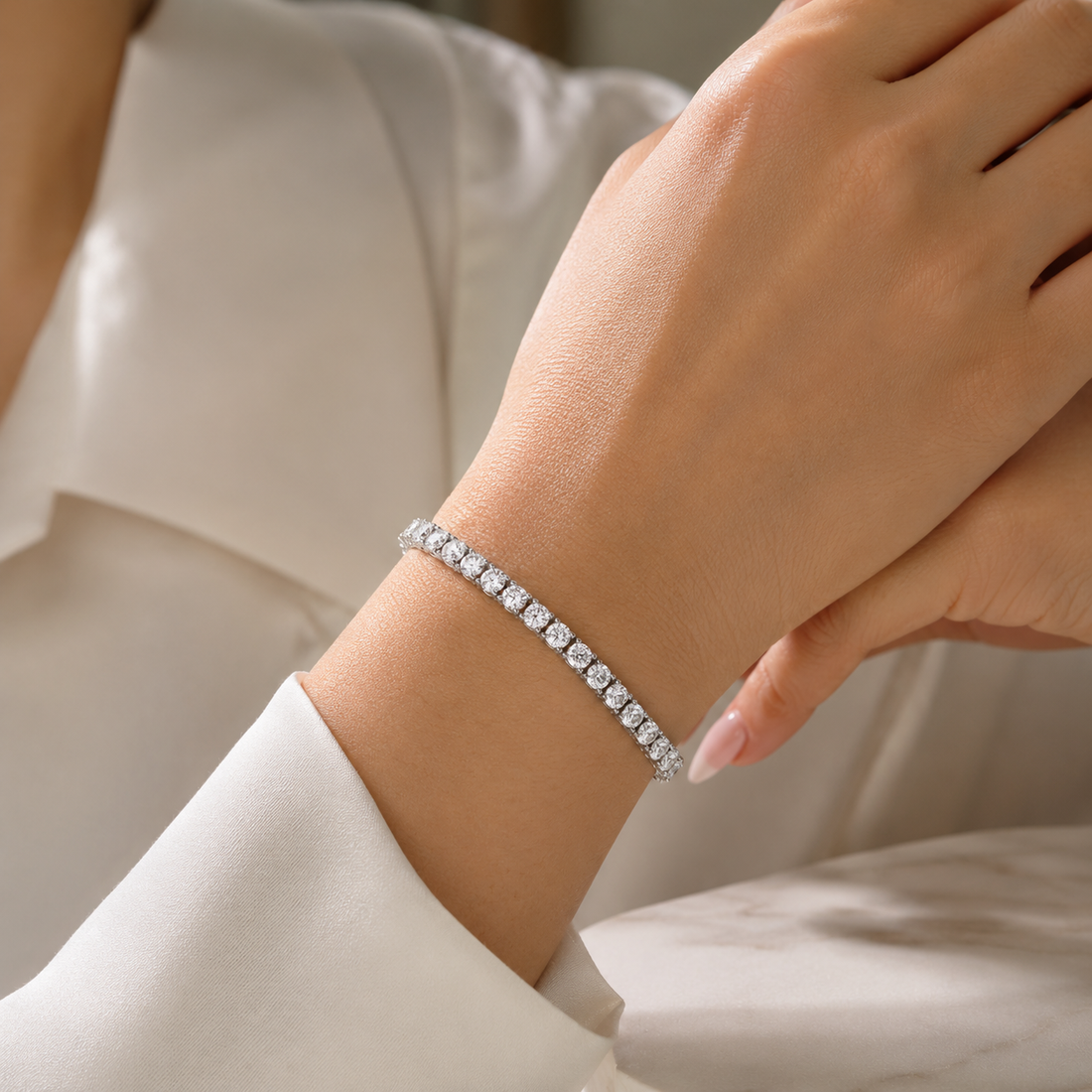 A close-up of a delicate silver tennis bracelet with clear stones worn on a woman's wrist, set against a soft white blouse.