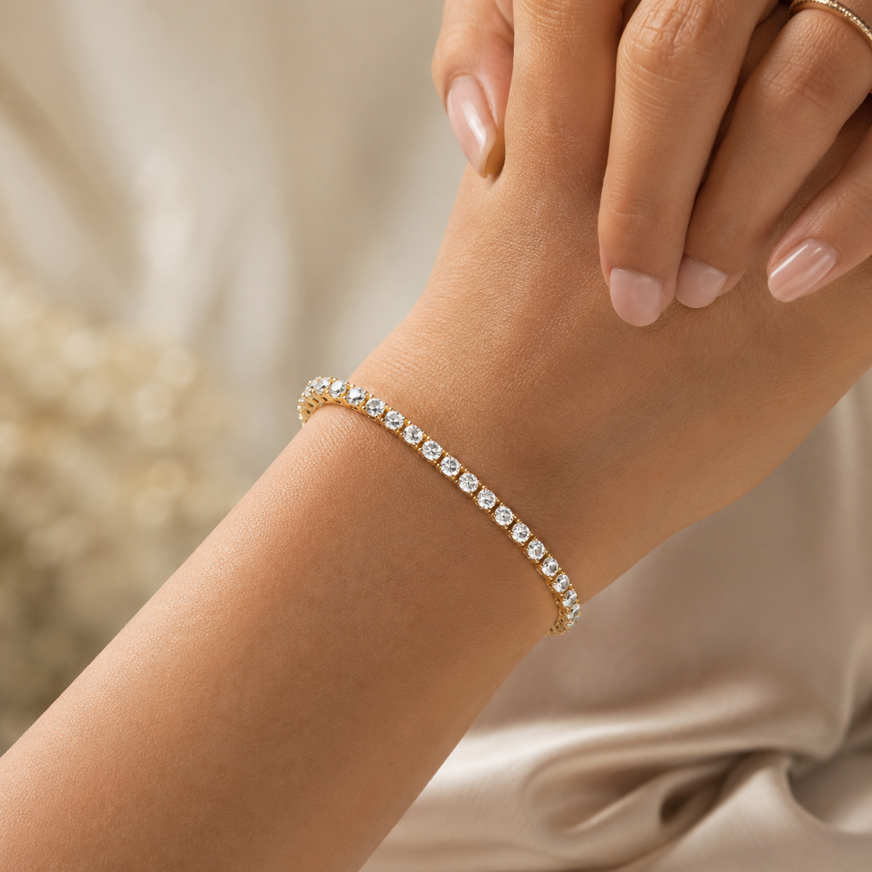 A delicate gold tennis bracelet with small clear stones, worn on a woman's wrist. The image is cropped to show the hand and wrist, with soft-focus beige fabric in the background.