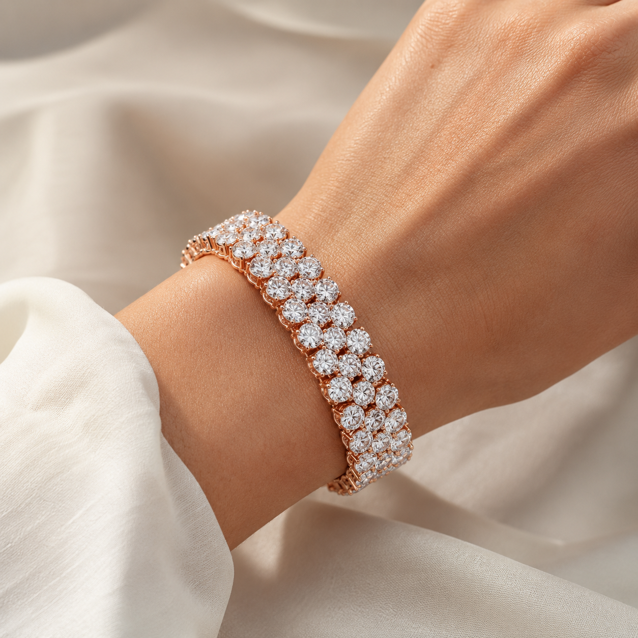 A close-up of a person's wrist wearing a rose gold bracelet adorned with multiple rows of sparkling clear gemstones, set against a soft, draped white fabric background.