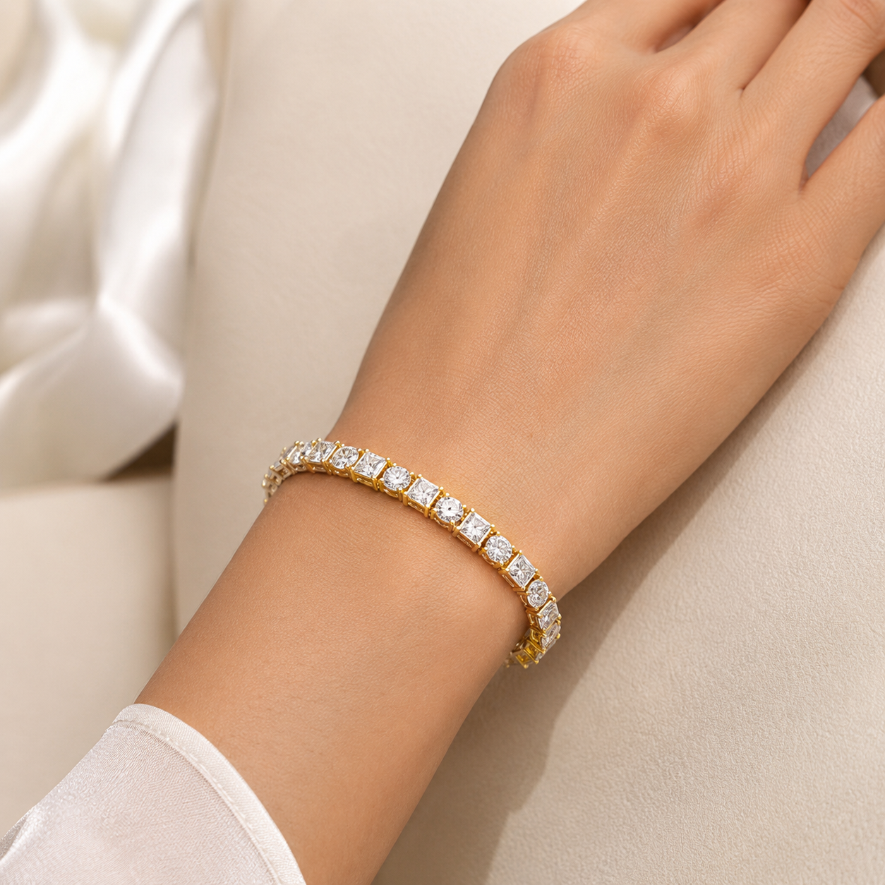 A close-up of a woman's wrist wearing a yellow gold tennis bracelet set with clear rectangular gemstones, resting against a soft beige background with white satin fabric.