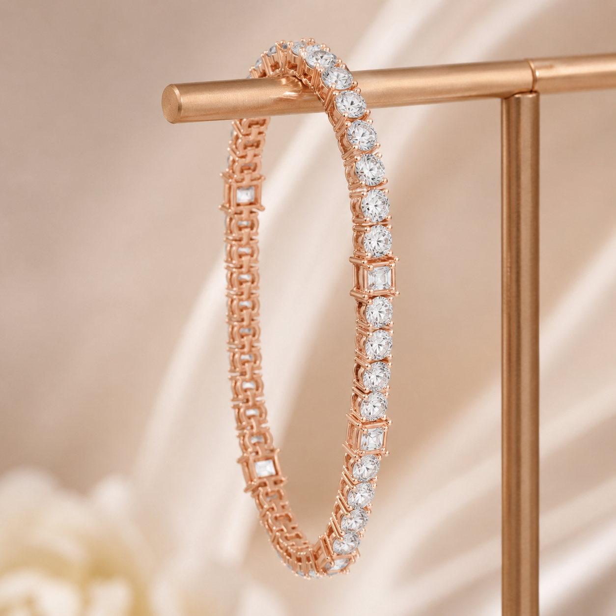 A rose gold tennis bracelet with clear rectangular and round stones, hanging from a rose gold jewelry stand against a soft beige satin background.