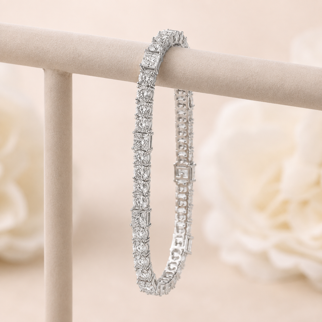 A silver tennis bracelet with square-cut clear stones, draped over a beige jewelry display stand against a soft, blurred cream background.