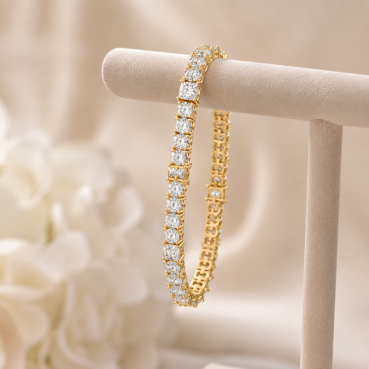 A gold tennis bracelet with rectangular clear stones, displayed on a cream jewelry stand against a soft beige background with blurred white flowers.