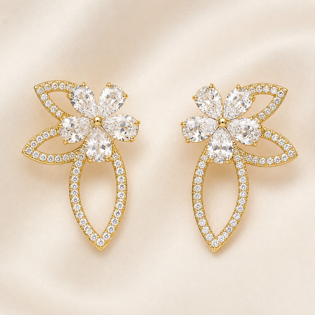 A pair of gold flower-shaped stud earrings adorned with clear marquise and round-cut stones, set against a soft beige satin background.