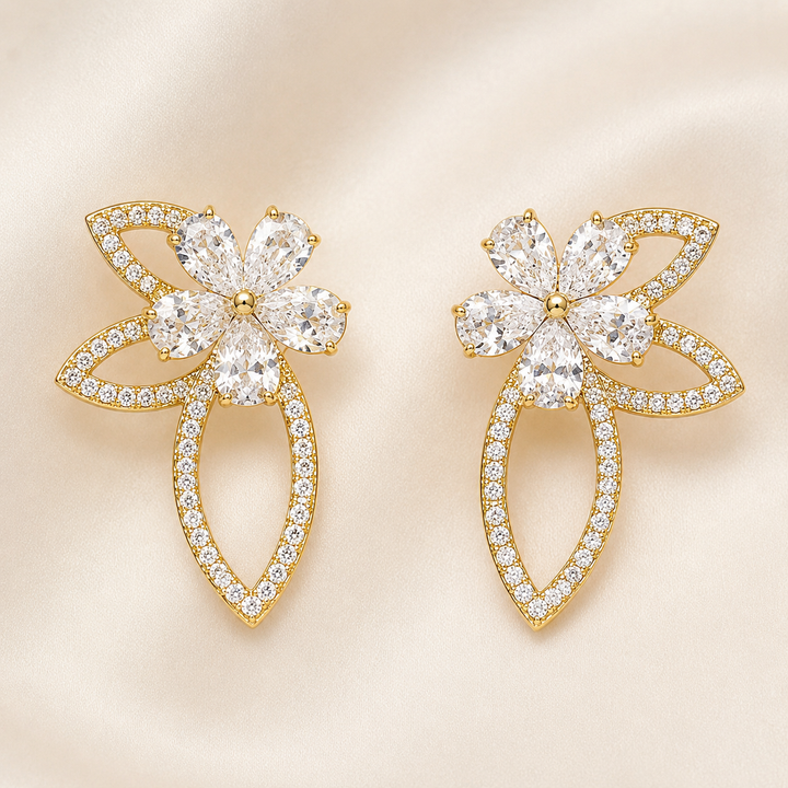 A pair of gold flower-shaped stud earrings adorned with clear marquise and round-cut stones, set against a soft beige satin background.
