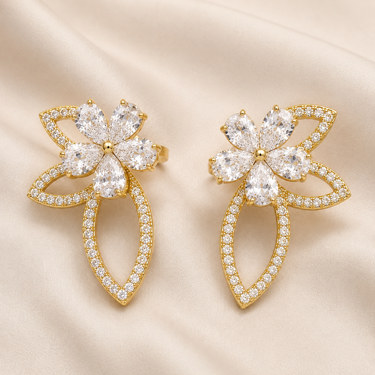 A pair of gold flower-shaped earrings adorned with clear crystals, displayed on a soft beige satin fabric background.
