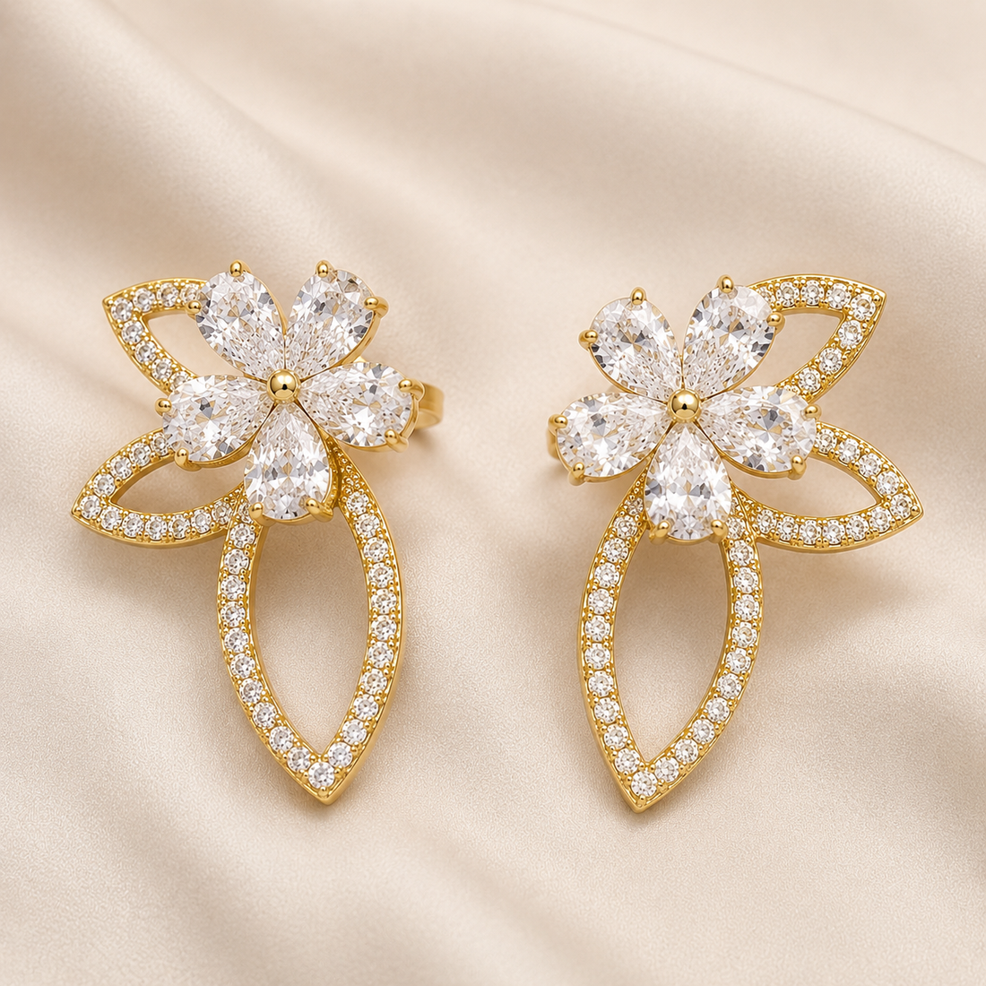 A pair of gold flower-shaped earrings adorned with clear crystals, displayed on a soft beige satin fabric background.