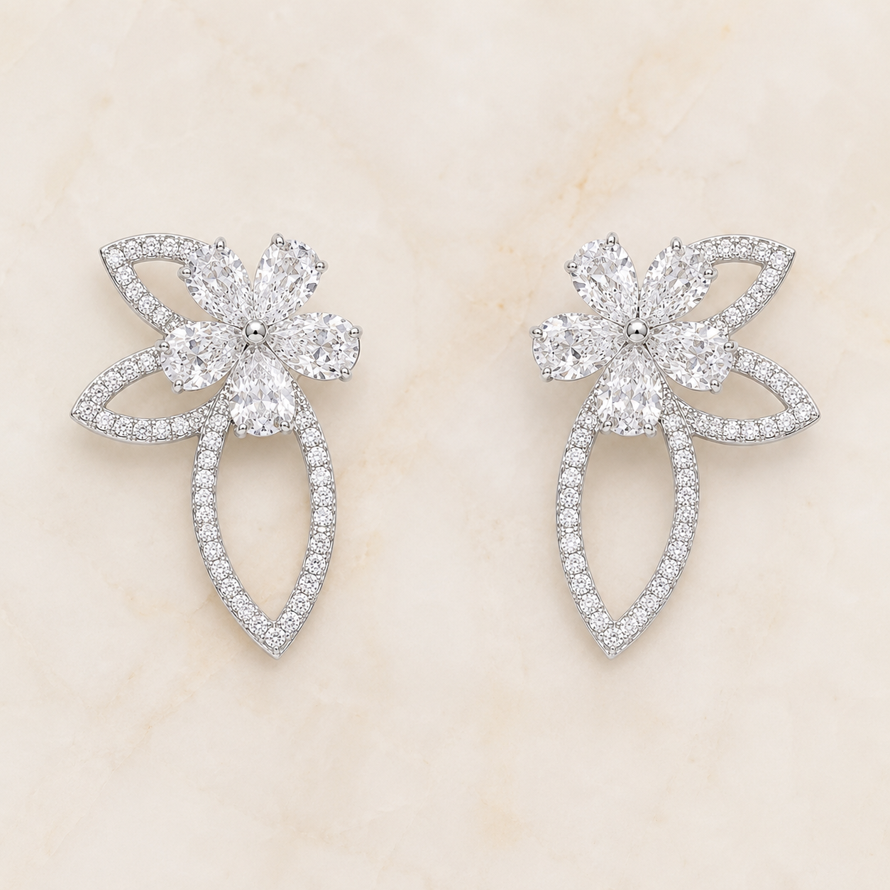 A pair of silver-toned flower-shaped earrings paved with clear round and marquise-cut gemstones, displayed on a light beige marble surface.