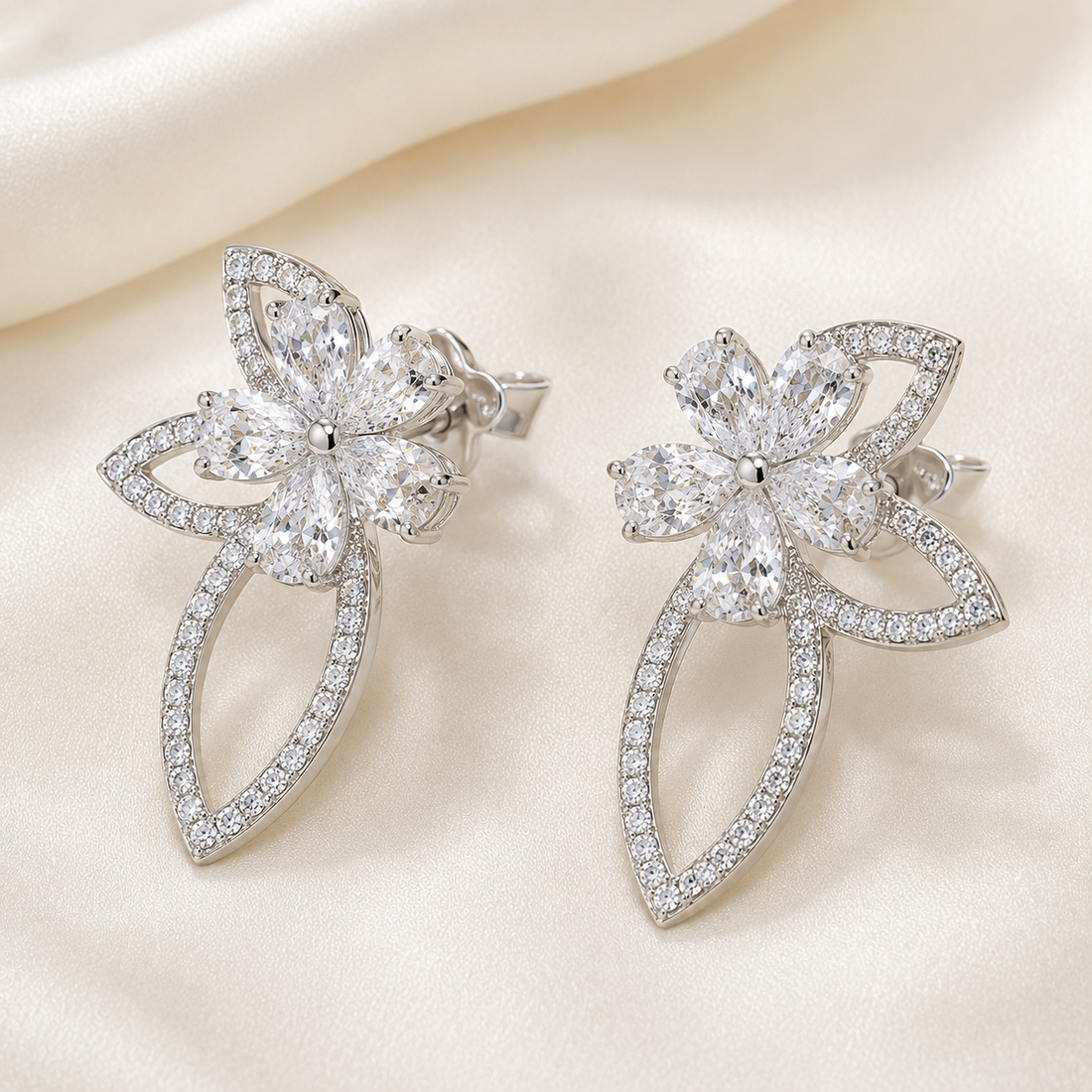 A pair of silver-toned flower-shaped earrings paved with clear cubic zirconia stones, displayed on a soft cream satin fabric background.