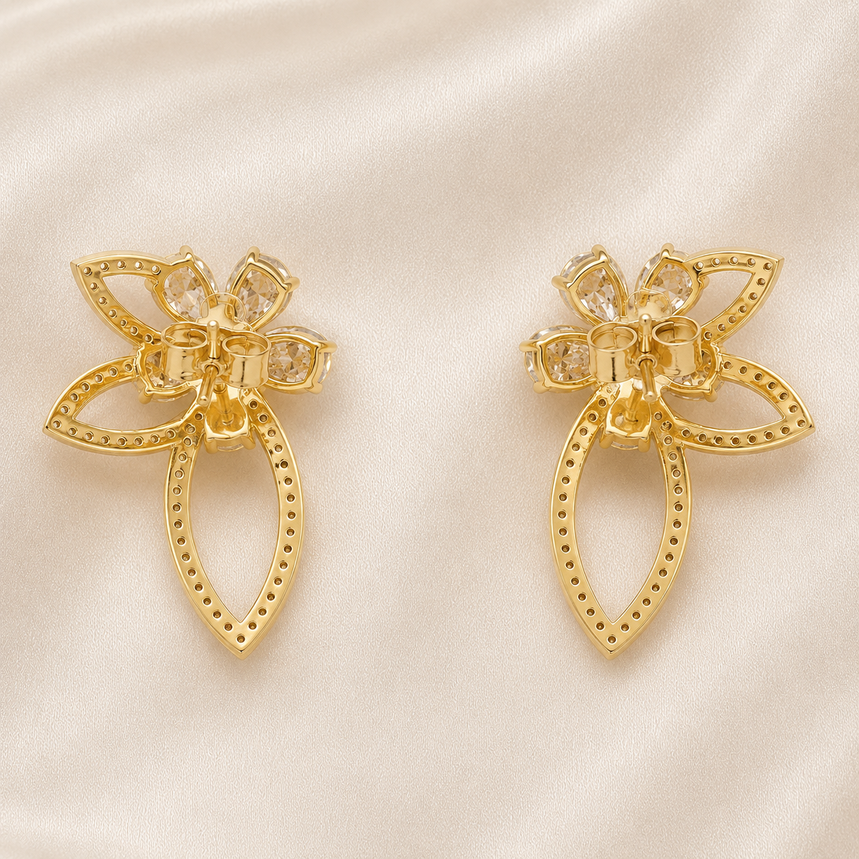 A pair of gold-toned flower-shaped earrings with clear rhinestone accents, displayed on a soft beige satin fabric background.