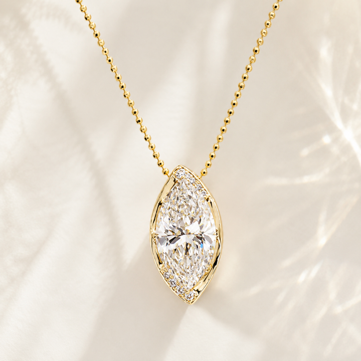 A gold necklace with a marquise-shaped diamond pendant, displayed on a soft, light-colored fabric background with delicate shadows.