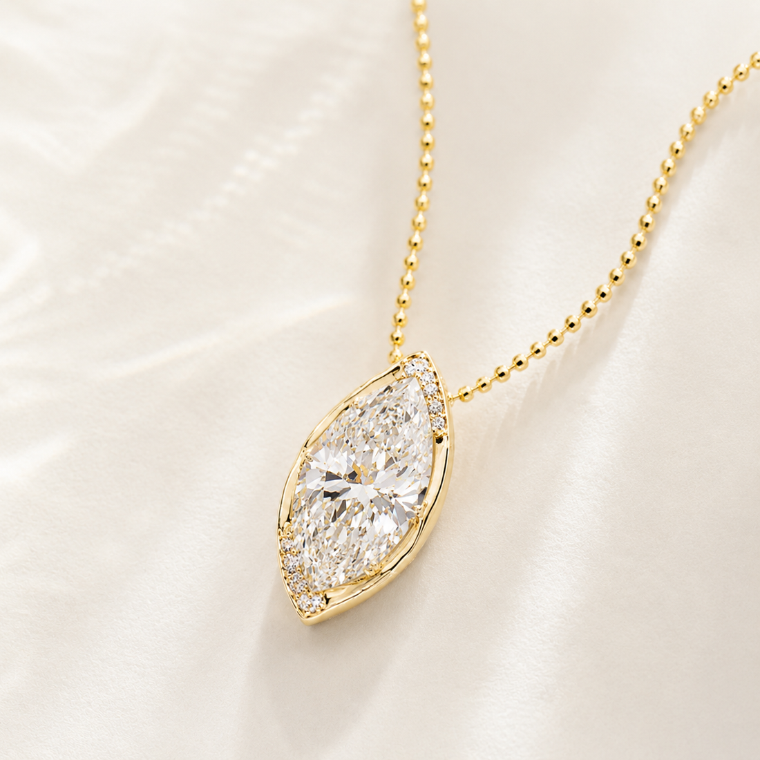 A gold necklace with a marquise-cut diamond pendant and a halo of small diamonds, displayed on a soft white satin fabric.