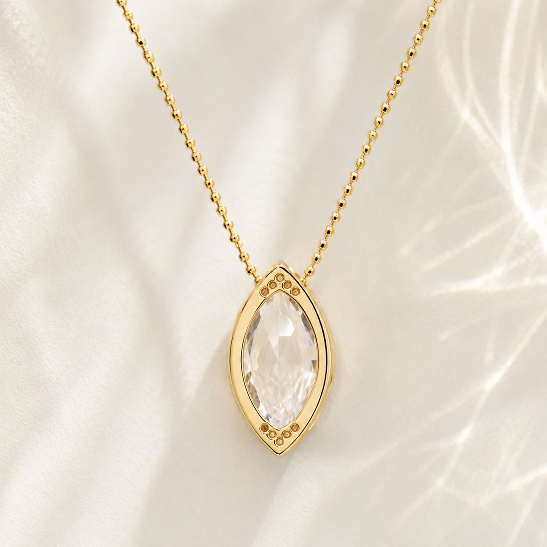 Gold necklace with a marquise-cut clear gemstone pendant, displayed on a soft white fabric background with delicate shadows.