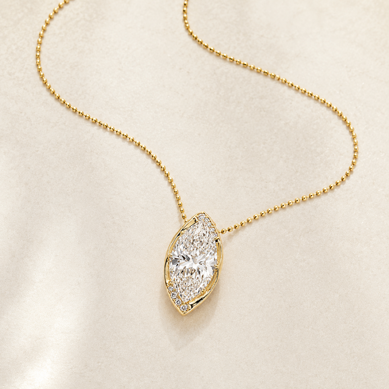 A gold necklace with a marquise-shaped pendant set with clear gemstones, displayed on a soft, light beige fabric background.