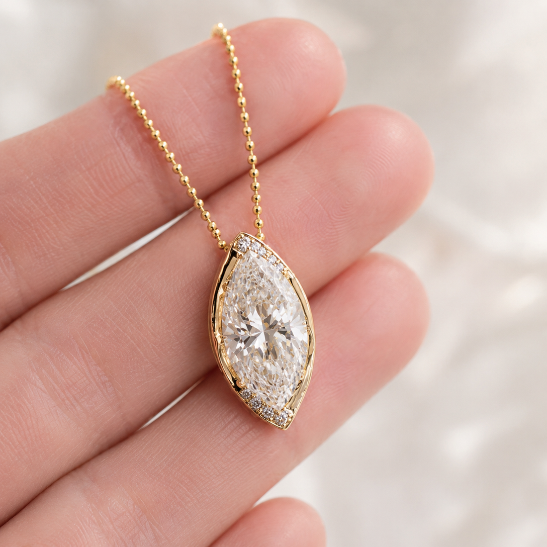 A gold pendant necklace with a marquise-cut clear gemstone and small accent stones, held on a person's fingers against a soft light background.