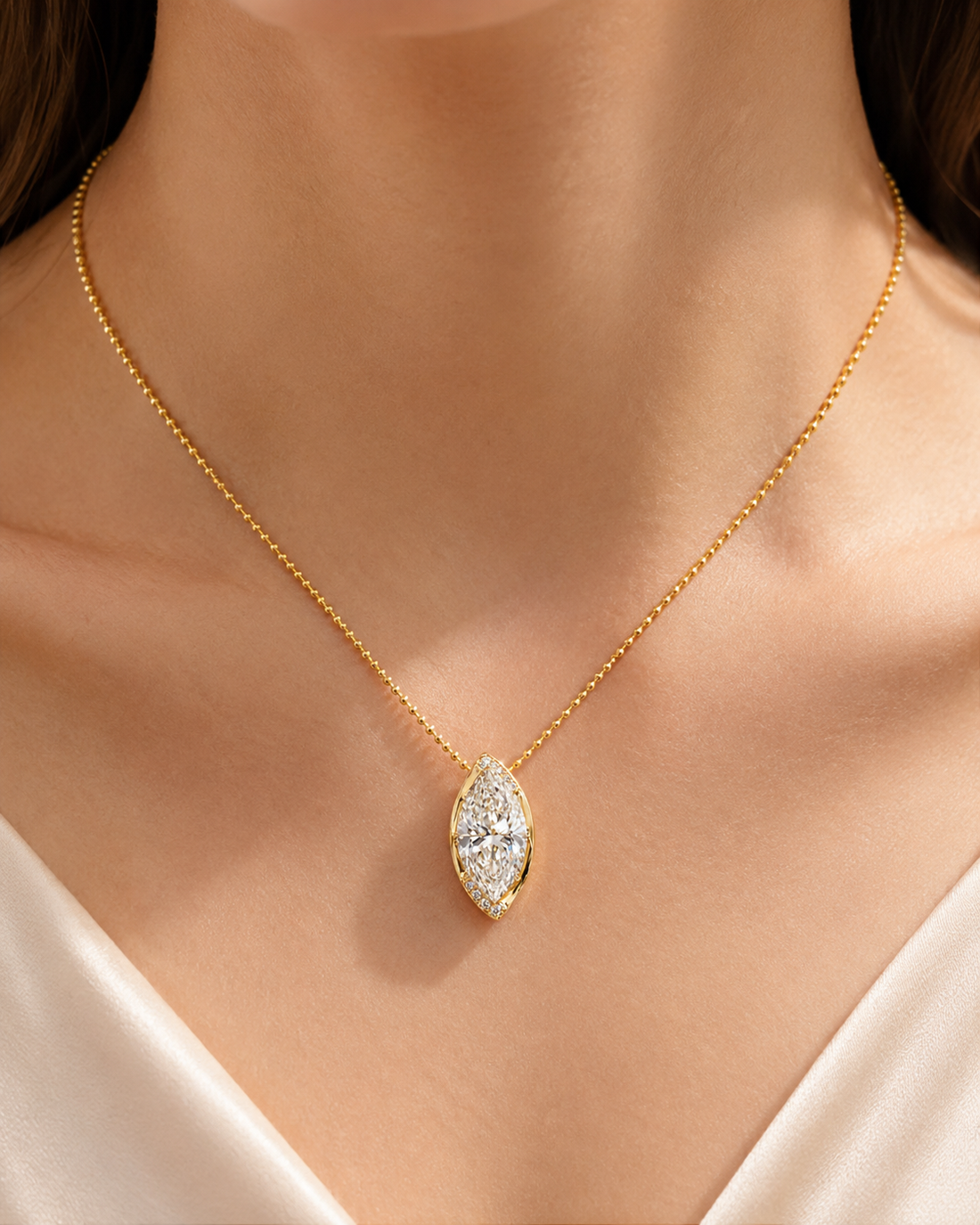 A close-up of a model's neck and chest wearing a delicate gold chain necklace with a marquise-shaped pendant set with clear stones.
