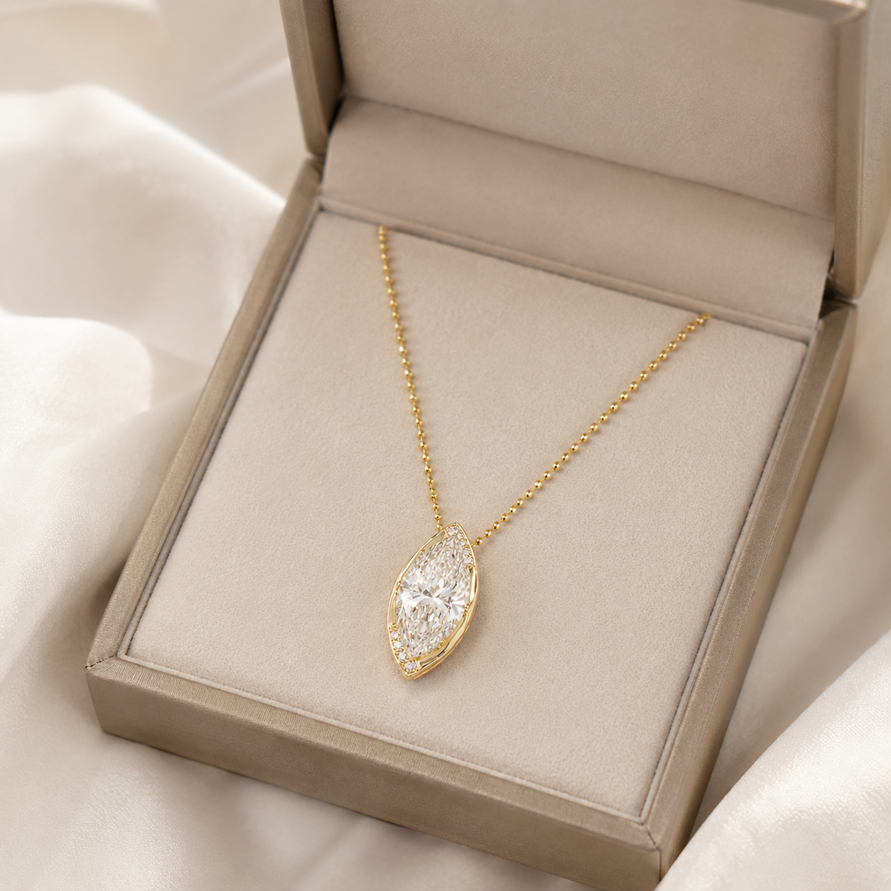 A gold pendant necklace with a marquise-shaped clear gemstone pavé setting, displayed inside an open beige jewelry box on soft white fabric.
