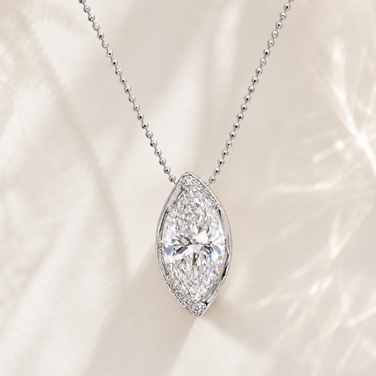 A silver-tone ball chain necklace with a marquise-shaped diamond pendant, accented by a halo of small round diamonds, set against a soft cream-colored textured background.