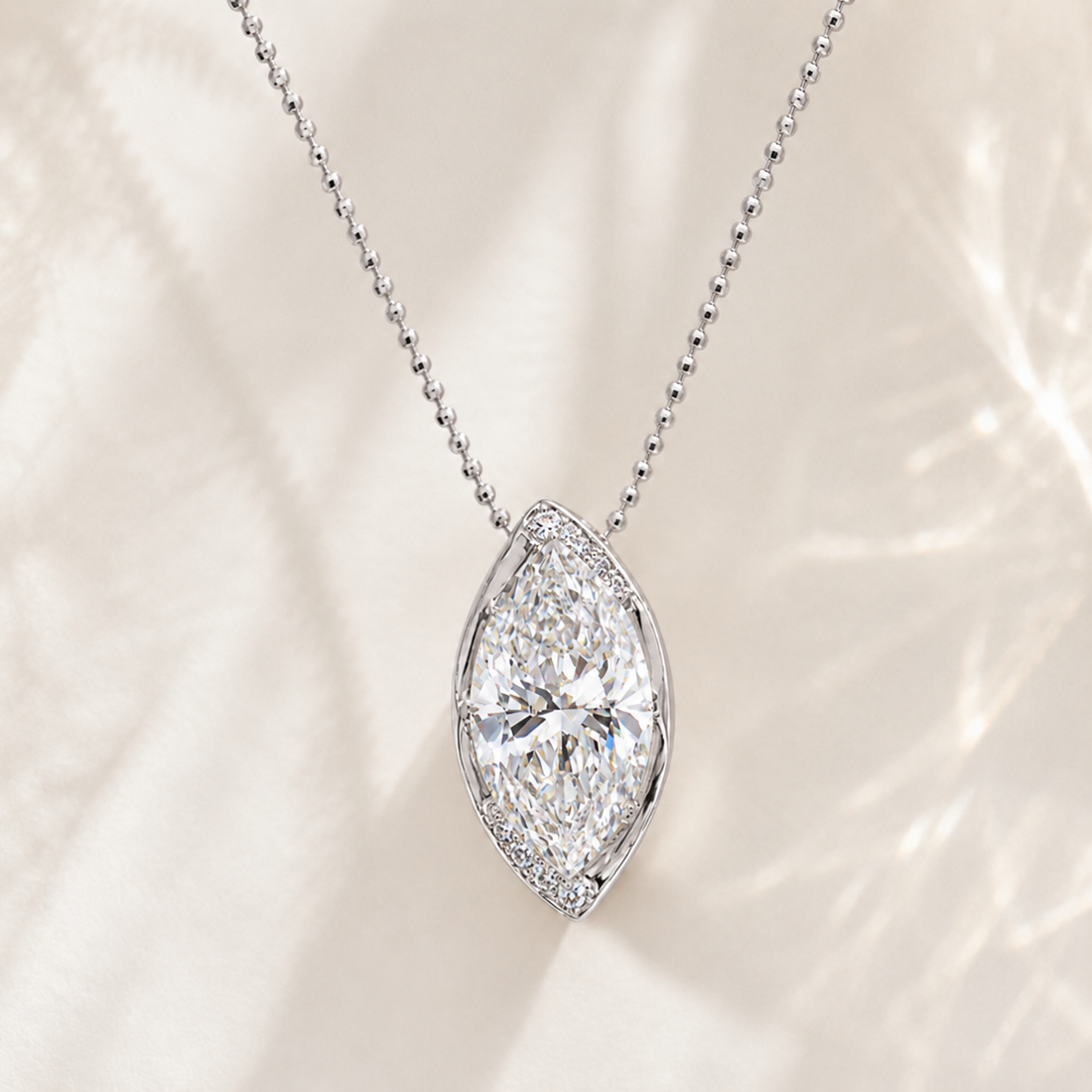 A silver-tone ball chain necklace with a marquise-shaped diamond pendant, accented by a halo of small round diamonds, set against a soft cream-colored textured background.