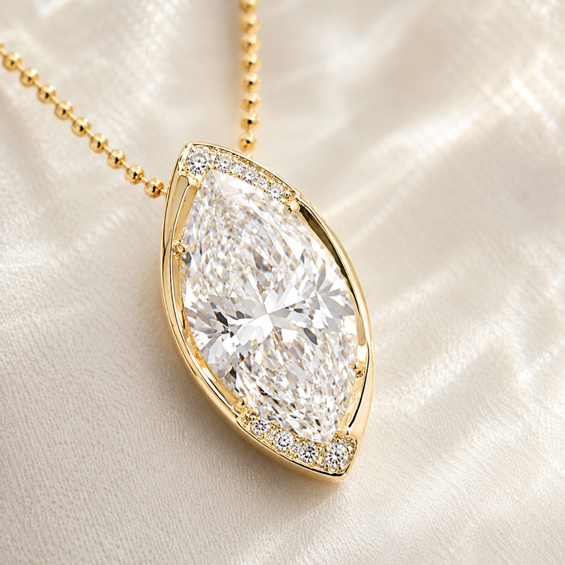 A gold pendant necklace featuring a large marquise-cut clear gemstone surrounded by small accent stones, resting on a soft white satin fabric background.