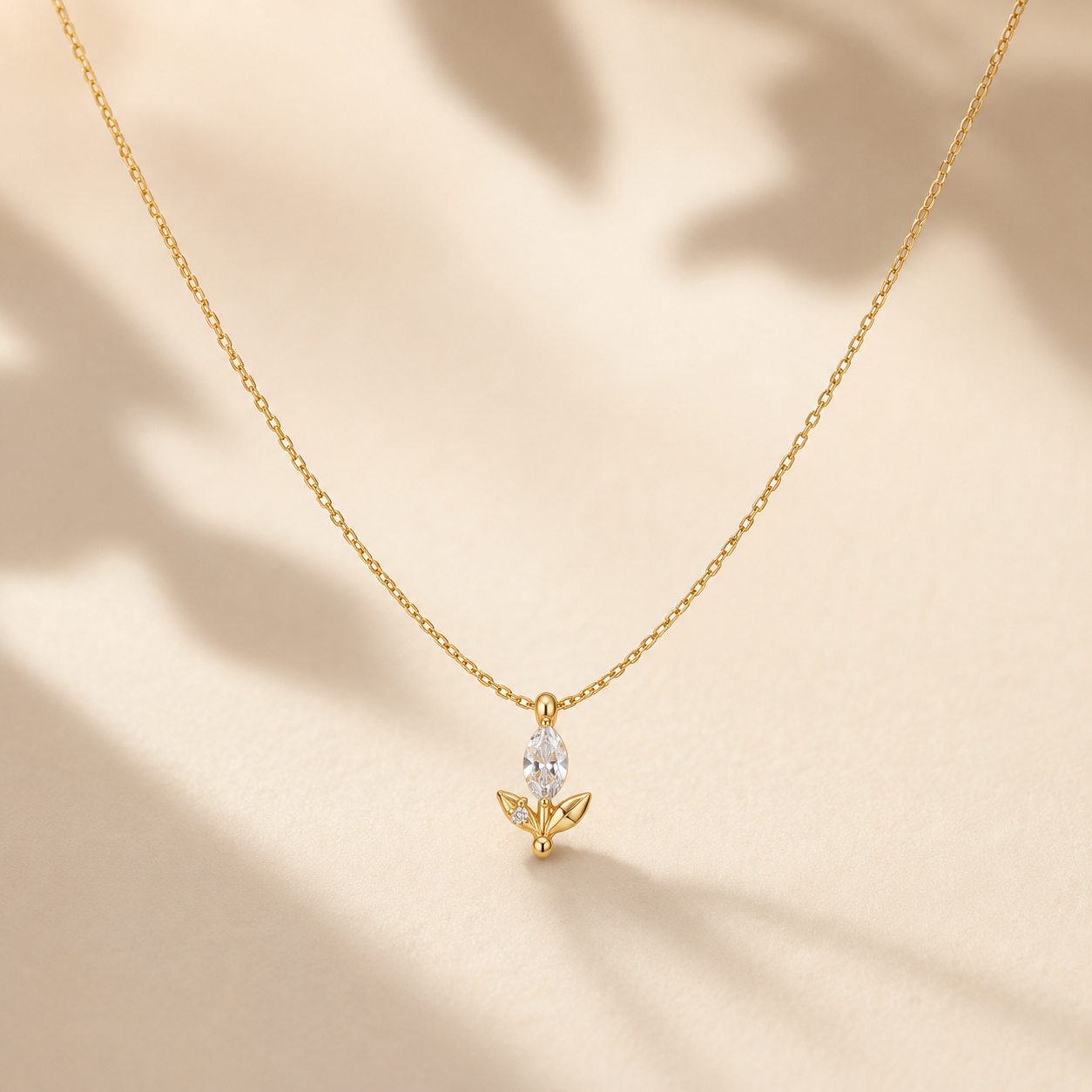 A delicate gold chain necklace with a small gold pendant featuring a marquise-cut clear gemstone and leaf-like details, displayed on a warm beige surface with soft leaf shadows.