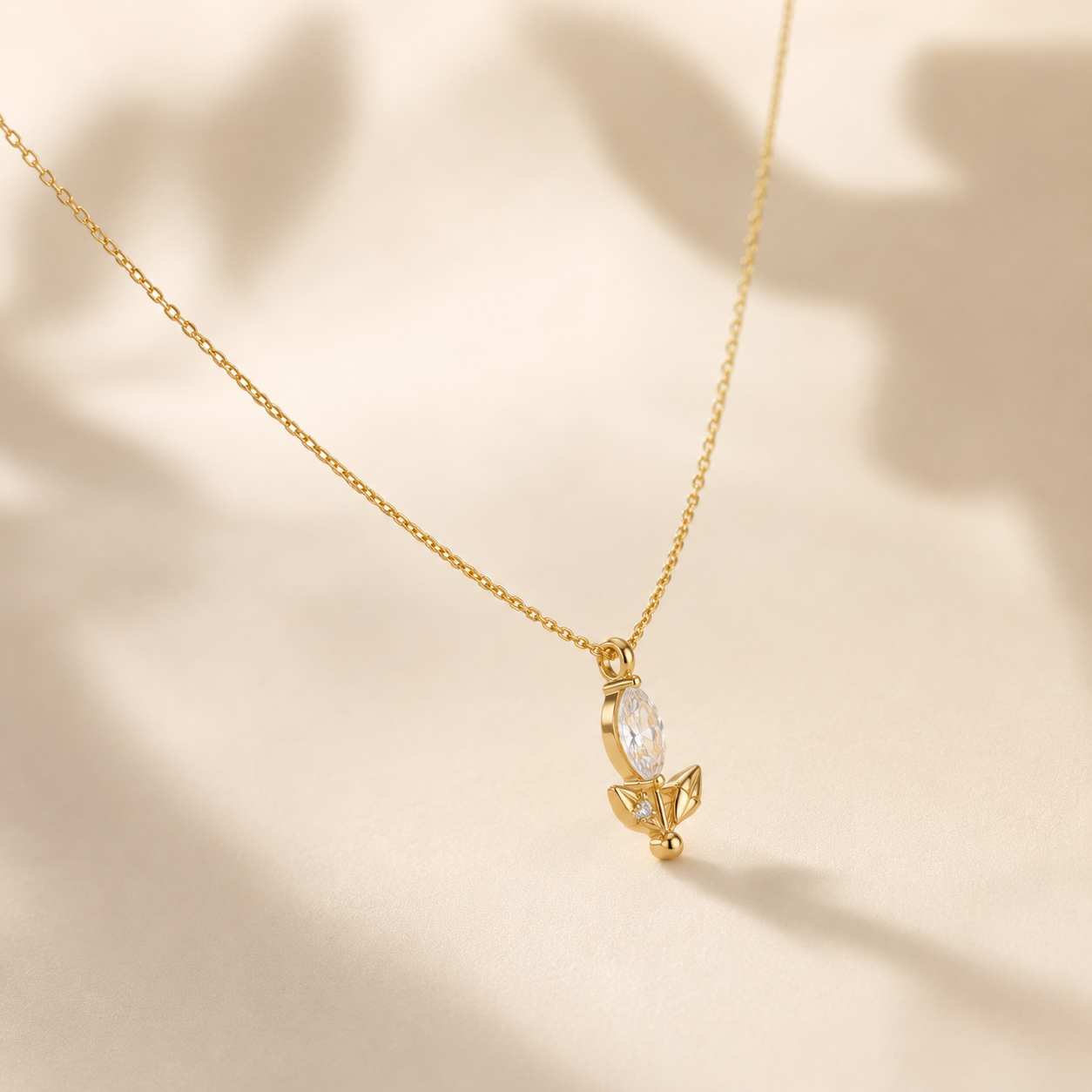 A delicate gold chain necklace with a small pendant featuring a marquise-cut clear stone and a gold leaf-like accent, displayed on a warm beige surface with soft leaf shadows.